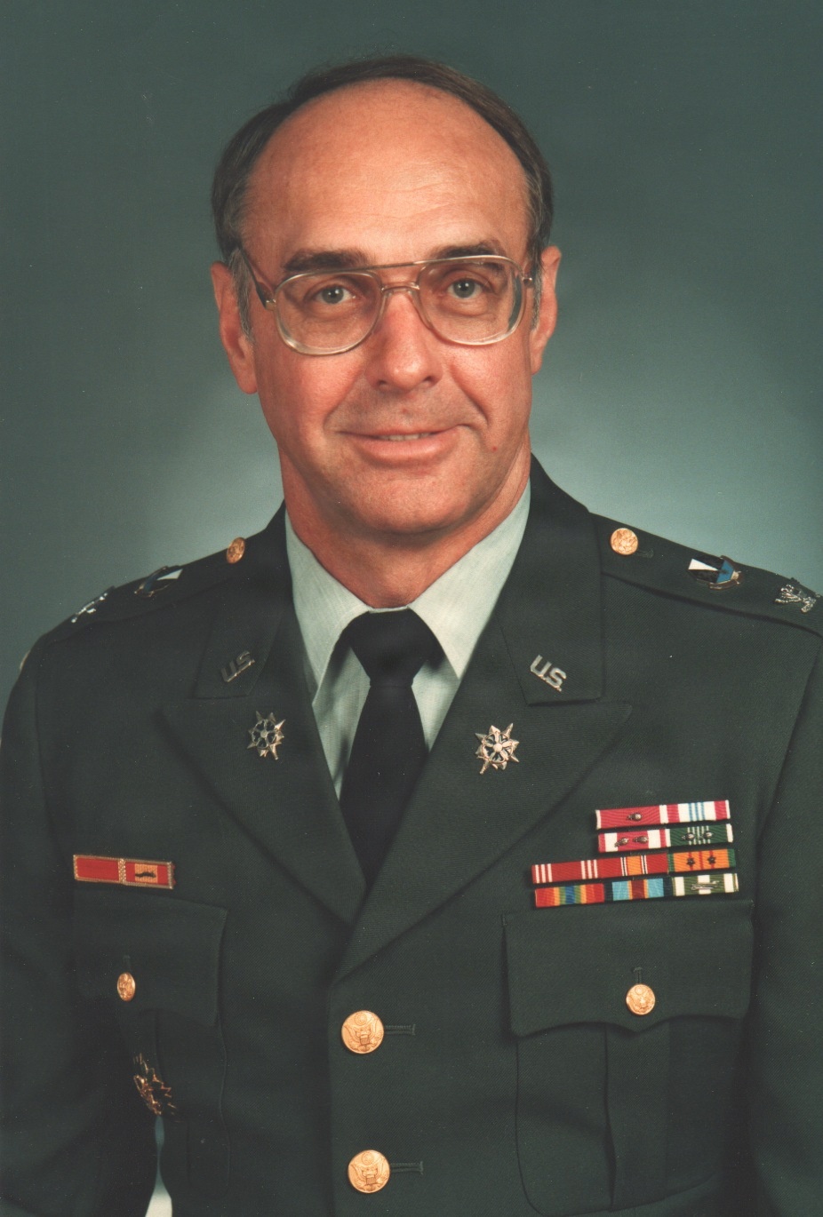 A formal military portrait of a U.S. Army officer in dress green uniform. The subject is a middle-aged male wearing glasses, displaying multiple ribbons, badges, and rank insignia consistent with a senior officer. The photo appears to be an official military portrait taken against a neutral studio background.
