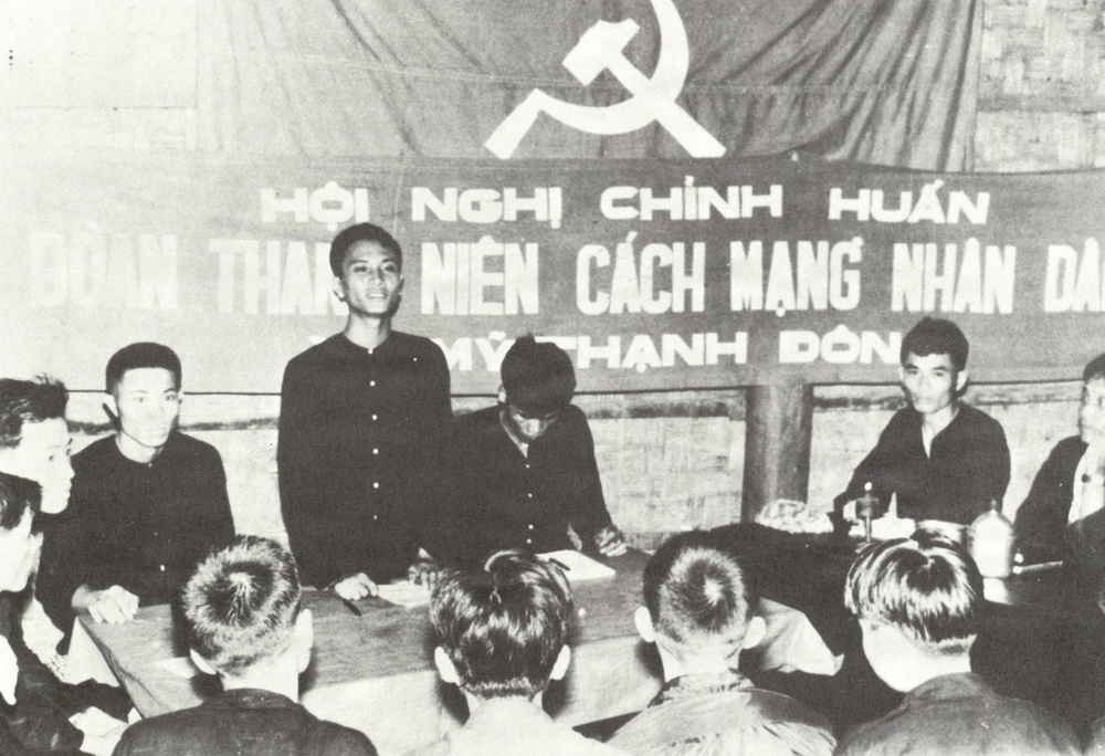 A black-and-white photograph showing a political meeting or indoctrination session held under a communist banner with hammer and sickle symbol. The banner text in Vietnamese reads 'Hội Nghị Chính Huấn' (Political Training Conference) and references 'Đoàn Thanh Niên Cách Mạng Nhân Dân' (People's Revolutionary Youth Union) at 'Mỹ Thạnh Đông'. Several young Vietnamese individuals are seated and standing around a table in what appears to be a formal organizational gathering.