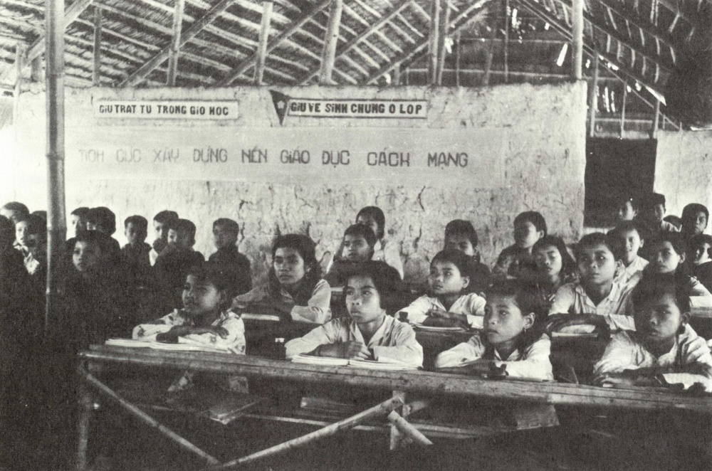 A black-and-white photograph of a crowded Vietnamese classroom, likely from the mid-20th century during the revolutionary period. Students of various ages sit at wooden desks in a rudimentary structure with a thatched roof and mud/plaster walls bearing Vietnamese-language slogans. The wall text references revolutionary education, consistent with North Vietnamese educational campaigns.