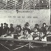 A black-and-white photograph of a crowded Vietnamese classroom, likely from the mid-20th century during the revolutionary period. Students of various ages sit at wooden desks in a rudimentary structure with a thatched roof and mud/plaster walls bearing Vietnamese-language slogans. The wall text references revolutionary education, consistent with North Vietnamese educational campaigns.