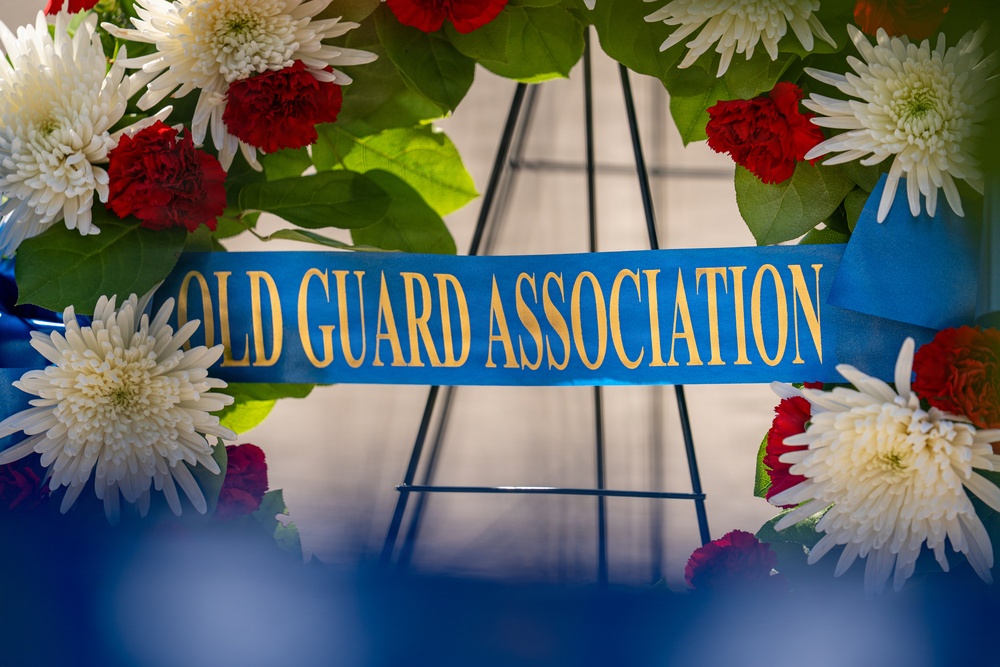 The image shows a formal memorial or ceremonial wreath adorned with red carnations and white chrysanthemums arranged in a circular pattern. A blue satin ribbon with gold lettering reads 'OLD GUARD ASSOCIATION,' indicating this wreath belongs to a military or veterans organization. The wreath is mounted on a metal stand, suggesting it is part of a formal commemorative ceremony.