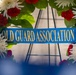 The image shows a formal memorial or ceremonial wreath adorned with red carnations and white chrysanthemums arranged in a circular pattern. A blue satin ribbon with gold lettering reads 'OLD GUARD ASSOCIATION,' indicating this wreath belongs to a military or veterans organization. The wreath is mounted on a metal stand, suggesting it is part of a formal commemorative ceremony.