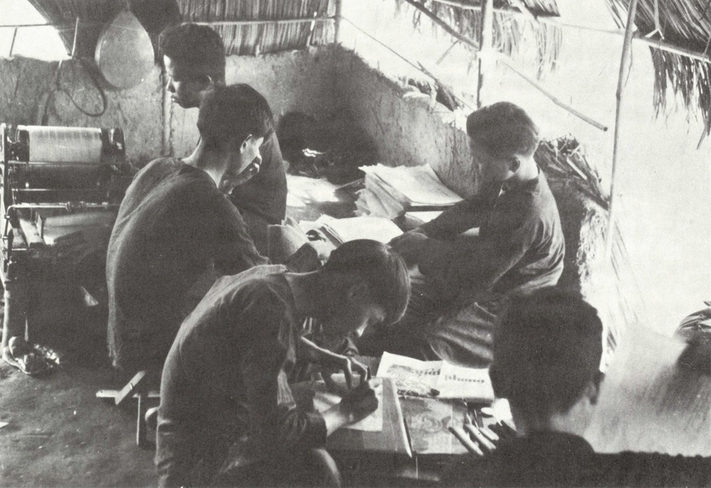 A black-and-white photograph showing a group of individuals gathered inside a rudimentary structure with a thatched roof and mud or clay walls. Several people appear to be engaged in writing, reading, or reviewing documents and printed materials at a shared workspace. The setting suggests a makeshift office, classroom, or editorial workspace in a rural or wartime environment.