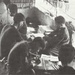 A black-and-white photograph showing a group of individuals gathered inside a rudimentary structure with a thatched roof and mud or clay walls. Several people appear to be engaged in writing, reading, or reviewing documents and printed materials at a shared workspace. The setting suggests a makeshift office, classroom, or editorial workspace in a rural or wartime environment.