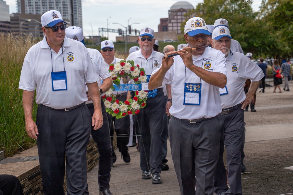A group of older men wearing matching white polo shirts and blue-and-white caps are participating in what appears to be a memorial procession or ceremony. They are carrying a floral wreath decorated with red and white flowers and a banner reading 'GUARD ASSOCIATION.' One member is playing a flute while walking, suggesting a formal commemorative event.