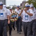 A group of older men wearing matching white polo shirts and blue-and-white caps are participating in what appears to be a memorial procession or ceremony. They are carrying a floral wreath decorated with red and white flowers and a banner reading 'GUARD ASSOCIATION.' One member is playing a flute while walking, suggesting a formal commemorative event.