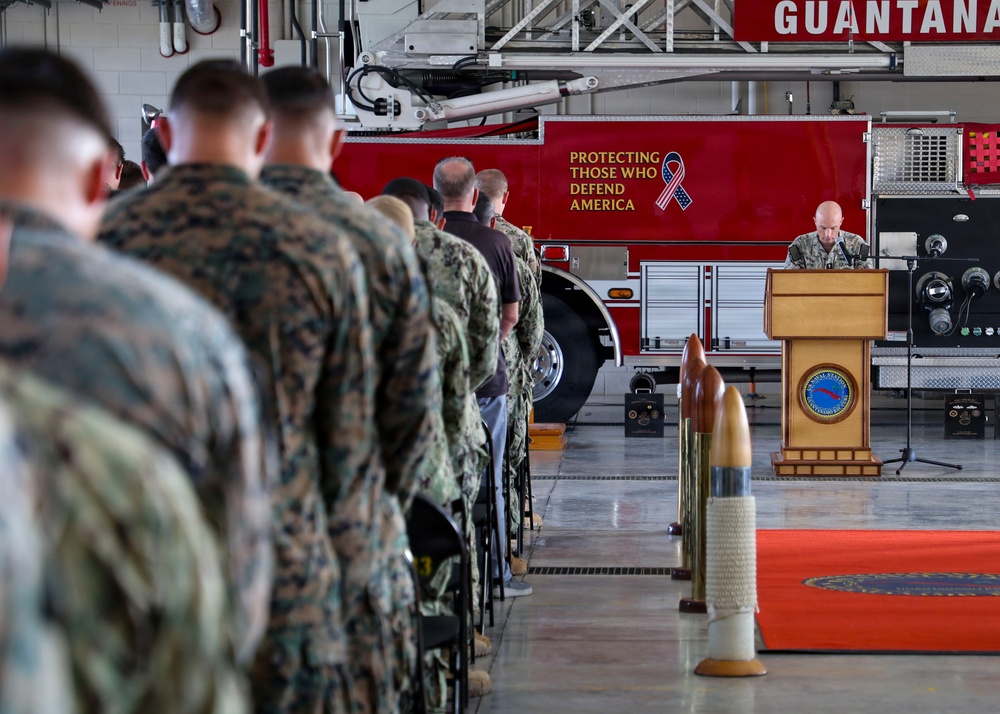 A military ceremony is taking place inside a fire station bay at Naval Station Guantanamo Bay. Service members in various uniforms, predominantly Marine Corps MARPAT camouflage, are standing in formation facing a speaker at a wooden podium. A red fire truck bearing the motto 'Protecting Those Who Defend America' serves as the backdrop.