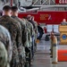 A military ceremony is taking place inside a fire station bay at Naval Station Guantanamo Bay. Service members in various uniforms, predominantly Marine Corps MARPAT camouflage, are standing in formation facing a speaker at a wooden podium. A red fire truck bearing the motto 'Protecting Those Who Defend America' serves as the backdrop.