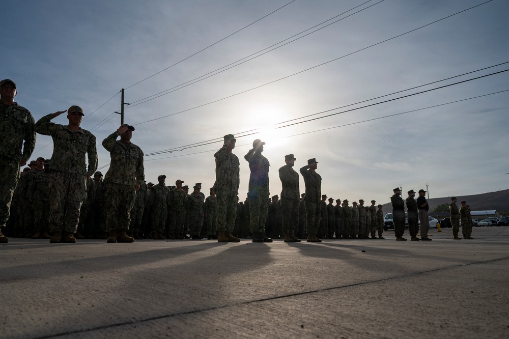 A large formation of military personnel in camouflage uniforms are standing at attention and rendering salutes on a paved surface, likely during a formal ceremony or morning colors. The image is taken against a bright backlit sun, creating strong silhouettes of the personnel. The scene appears to take place at a military installation, with utility poles, power lines, and buildings visible in the background.