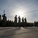 A large formation of military personnel in camouflage uniforms are standing at attention and rendering salutes on a paved surface, likely during a formal ceremony or morning colors. The image is taken against a bright backlit sun, creating strong silhouettes of the personnel. The scene appears to take place at a military installation, with utility poles, power lines, and buildings visible in the background.