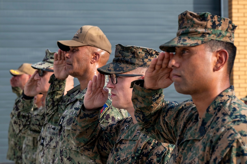 A group of military personnel in various camouflage uniforms are standing in formation and rendering a salute. The group includes members from multiple branches, visibly identified by 'U.S. Navy' and 'Marines' uniform markings. The scene appears to be an outdoor military ceremony or formal event.