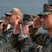 A group of military personnel in various camouflage uniforms are standing in formation and rendering a salute. The group includes members from multiple branches, visibly identified by 'U.S. Navy' and 'Marines' uniform markings. The scene appears to be an outdoor military ceremony or formal event.