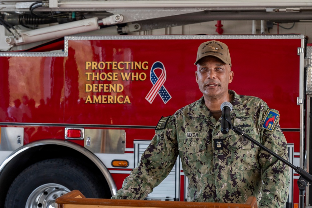 A U.S. Navy officer with the name tag 'NIXON' is speaking at a podium in front of a red fire truck at what appears to be U.S. Naval Station Guantanamo Bay, Cuba. The officer is wearing Navy Working Uniform (NWU) camouflage and a tan cap with 'U.S. Naval Station Guantanamo Bay, Cuba' text. The fire truck bears the inscription 'Protecting Those Who Defend America' alongside an American flag ribbon.