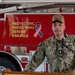 A U.S. Navy officer with the name tag 'NIXON' is speaking at a podium in front of a red fire truck at what appears to be U.S. Naval Station Guantanamo Bay, Cuba. The officer is wearing Navy Working Uniform (NWU) camouflage and a tan cap with 'U.S. Naval Station Guantanamo Bay, Cuba' text. The fire truck bears the inscription 'Protecting Those Who Defend America' alongside an American flag ribbon.