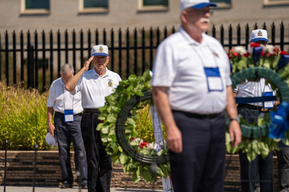 A solemn memorial ceremony is taking place outdoors, featuring several older men dressed in matching white polo shirts and white-and-blue caps. One man is rendering a formal military salute while others stand at attention near decorative wreaths adorned with red, white, and blue ribbons. The setting appears to be a formal commemorative event, likely a veterans or military memorial service.