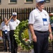 A solemn memorial ceremony is taking place outdoors, featuring several older men dressed in matching white polo shirts and white-and-blue caps. One man is rendering a formal military salute while others stand at attention near decorative wreaths adorned with red, white, and blue ribbons. The setting appears to be a formal commemorative event, likely a veterans or military memorial service.