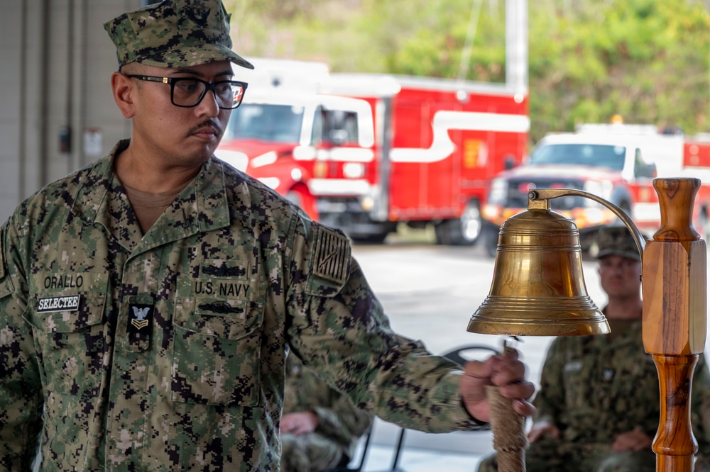 A U.S. Navy sailor identified as 'Orallo' with a 'SELECTEE' tag is ringing a brass ship's bell mounted on a wooden stand during what appears to be a military ceremony. The sailor is wearing Navy Working Uniform (NWU) digital camouflage and holds a rope attached to the bell's clapper. The setting appears to be near a fire station or vehicle bay, with red fire trucks visible in the background.