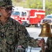 A U.S. Navy sailor identified as 'Orallo' with a 'SELECTEE' tag is ringing a brass ship's bell mounted on a wooden stand during what appears to be a military ceremony. The sailor is wearing Navy Working Uniform (NWU) digital camouflage and holds a rope attached to the bell's clapper. The setting appears to be near a fire station or vehicle bay, with red fire trucks visible in the background.