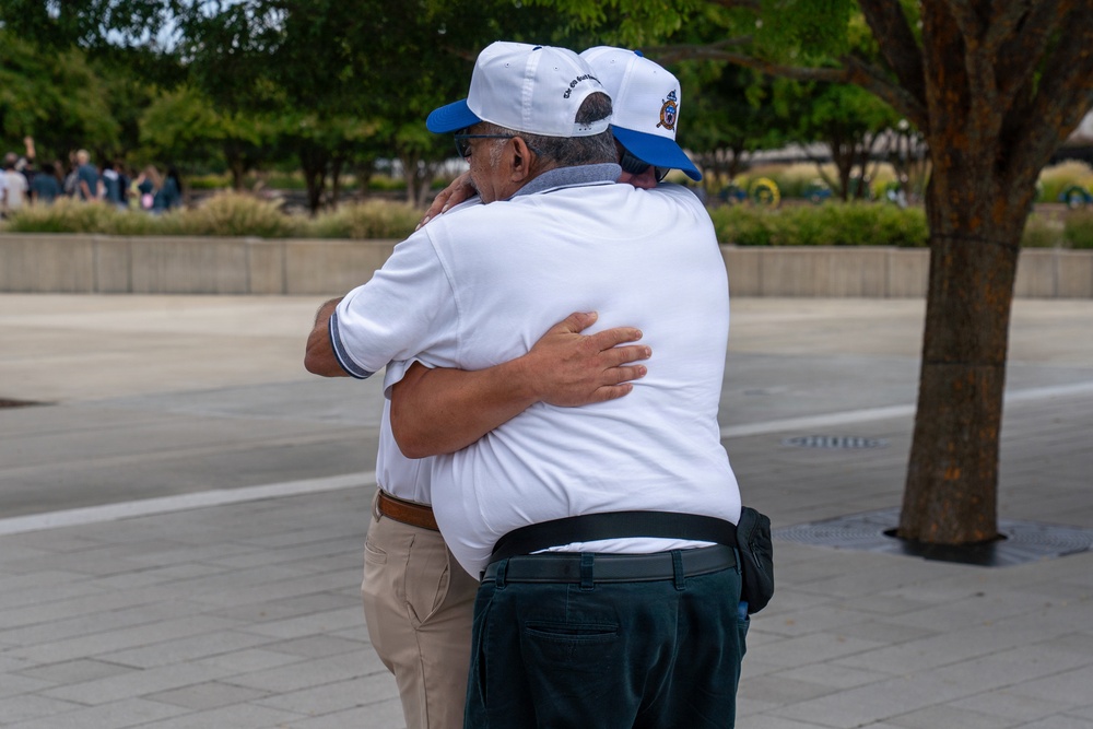 Two older men are embracing in what appears to be an outdoor public plaza or memorial space. Both are wearing matching white caps with blue brims that appear to have insignia or text on them, and white polo shirts, suggesting they may be part of the same organization or group. The emotional hug suggests a meaningful reunion or shared experience.