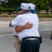 Two older men are embracing in what appears to be an outdoor public plaza or memorial space. Both are wearing matching white caps with blue brims that appear to have insignia or text on them, and white polo shirts, suggesting they may be part of the same organization or group. The emotional hug suggests a meaningful reunion or shared experience.