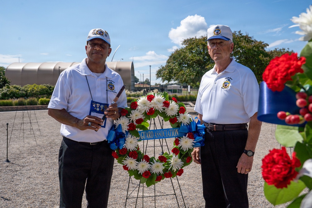Two men wearing matching white polo shirts and caps with an organizational emblem stand beside a memorial wreath at an outdoor ceremony. The wreath, composed of red and white flowers with a blue ribbon reading 'GUARD ASSOCIATION,' is displayed on a stand, suggesting a commemorative or veterans' event. The setting appears to be a memorial or public grounds on a clear, sunny day.