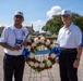 Two men wearing matching white polo shirts and caps with an organizational emblem stand beside a memorial wreath at an outdoor ceremony. The wreath, composed of red and white flowers with a blue ribbon reading 'GUARD ASSOCIATION,' is displayed on a stand, suggesting a commemorative or veterans' event. The setting appears to be a memorial or public grounds on a clear, sunny day.