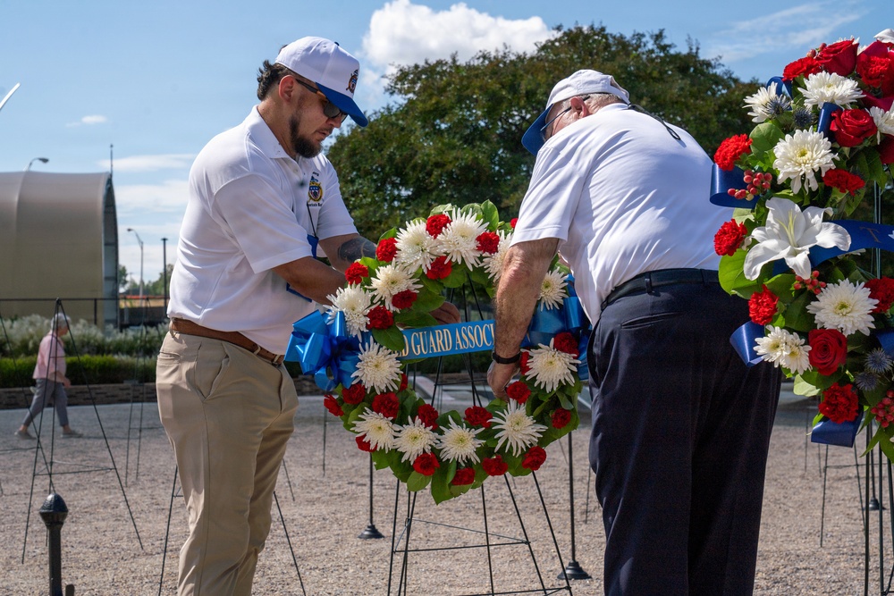 Two men dressed in white shirts and caps are arranging a memorial wreath at what appears to be a commemorative ceremony. The wreath bears a blue ribbon with text reading 'GUARD ASSOC' (partially visible), suggesting affiliation with a Guard Association. Multiple floral wreaths with red, white, and blue flowers are displayed on stands in an outdoor plaza setting.