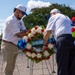 Two men dressed in white shirts and caps are arranging a memorial wreath at what appears to be a commemorative ceremony. The wreath bears a blue ribbon with text reading 'GUARD ASSOC' (partially visible), suggesting affiliation with a Guard Association. Multiple floral wreaths with red, white, and blue flowers are displayed on stands in an outdoor plaza setting.