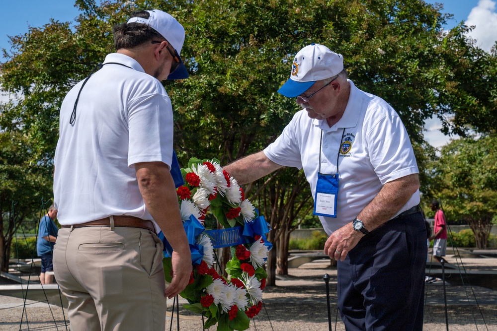 Two men in white polo shirts and caps are placing or adjusting a red, white, and blue floral wreath at what appears to be a memorial ceremony. The wreath features a blue ribbon with text indicating an association affiliation. The setting appears to be an outdoor memorial site with manicured grounds and trees in the background.