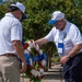 Two men in white polo shirts and caps are placing or adjusting a red, white, and blue floral wreath at what appears to be a memorial ceremony. The wreath features a blue ribbon with text indicating an association affiliation. The setting appears to be an outdoor memorial site with manicured grounds and trees in the background.