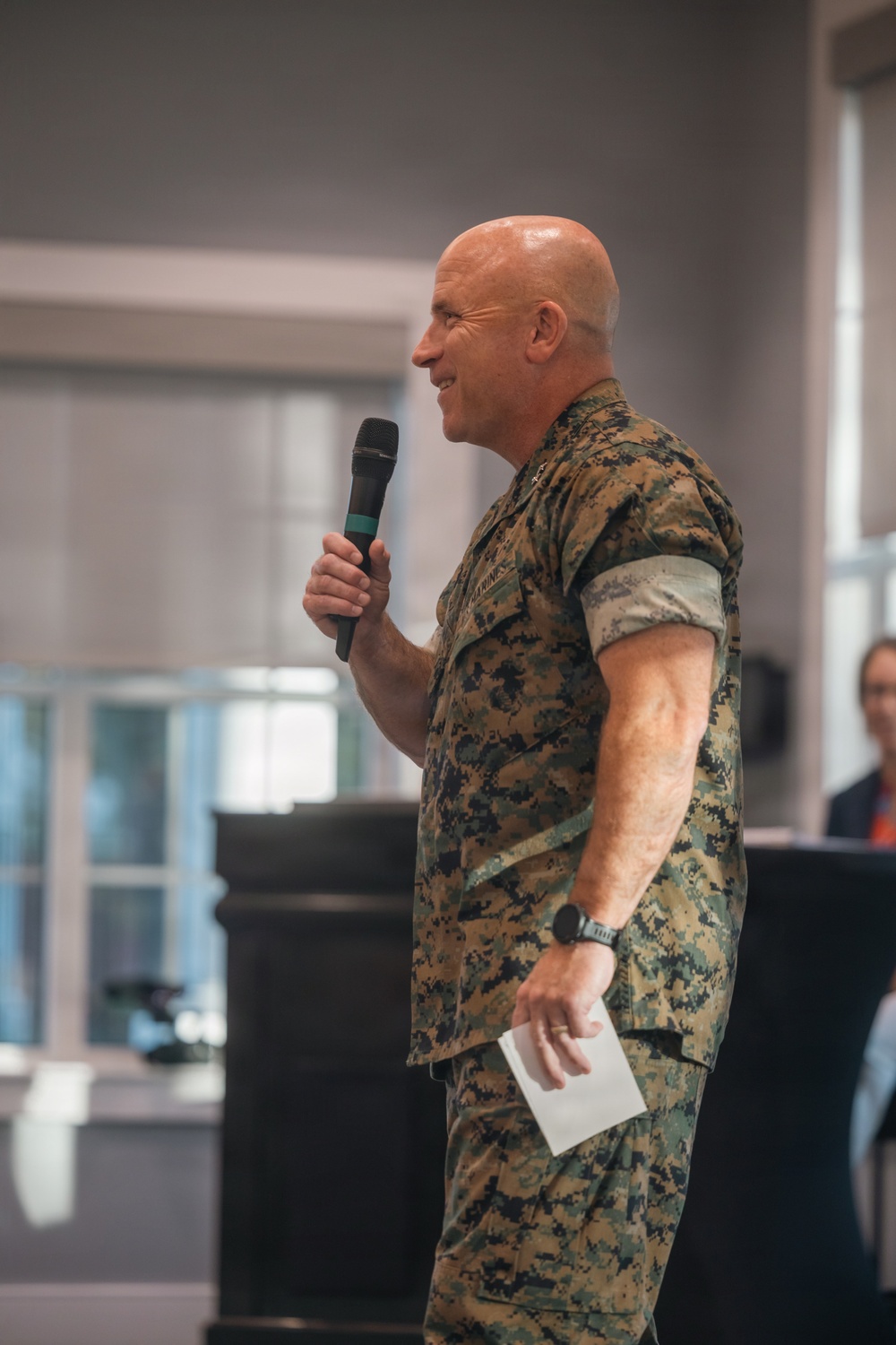 A bald male individual wearing U.S. Marine Corps MARPAT woodland camouflage uniform is speaking into a handheld microphone at what appears to be an indoor event or briefing. He is holding a small piece of paper in his other hand and appears to be smiling, suggesting a formal or semi-formal speaking engagement. The background shows a well-lit interior space with windows and at least one other person partially visible.