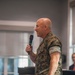A bald male individual wearing U.S. Marine Corps MARPAT woodland camouflage uniform is speaking into a handheld microphone at what appears to be an indoor event or briefing. He is holding a small piece of paper in his other hand and appears to be smiling, suggesting a formal or semi-formal speaking engagement. The background shows a well-lit interior space with windows and at least one other person partially visible.