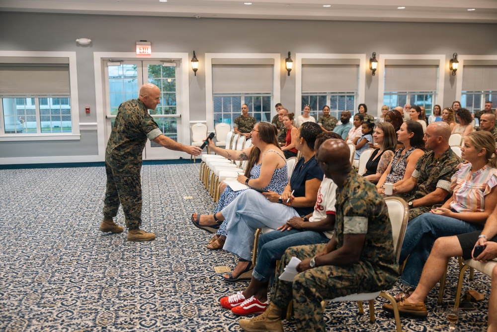 A U.S. Marine in camouflage uniform is conducting a town hall or briefing session, extending a microphone toward a seated audience member in a formal meeting room. The audience consists of a mix of uniformed Marines and civilian attendees, likely military family members or community members. The setting appears to be a military installation community center or club facility.