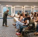 A U.S. Marine in camouflage uniform is conducting a town hall or briefing session, extending a microphone toward a seated audience member in a formal meeting room. The audience consists of a mix of uniformed Marines and civilian attendees, likely military family members or community members. The setting appears to be a military installation community center or club facility.