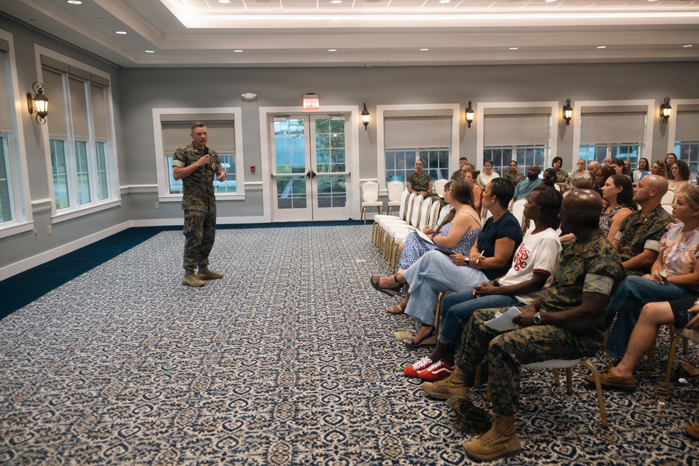 A U.S. Marine in camouflage uniform is addressing a seated audience in a formal banquet or conference room. The audience consists of a mix of civilians and uniformed Marines, suggesting a military briefing or family readiness event. The setting appears to be a well-appointed facility, likely on a military installation.