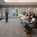 A U.S. Marine in camouflage uniform is addressing a seated audience in a formal banquet or conference room. The audience consists of a mix of civilians and uniformed Marines, suggesting a military briefing or family readiness event. The setting appears to be a well-appointed facility, likely on a military installation.
