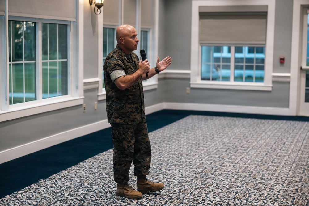 A bald male individual wearing U.S. Marine Corps MARPAT camouflage uniform is standing in what appears to be a formal event hall or conference room, speaking into a handheld microphone. He is gesturing with his free hand, suggesting he is delivering a speech or briefing. The room features large windows, decorative patterned carpet, and neutral-toned walls.