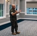 A bald male individual wearing U.S. Marine Corps MARPAT camouflage uniform is standing in what appears to be a formal event hall or conference room, speaking into a handheld microphone. He is gesturing with his free hand, suggesting he is delivering a speech or briefing. The room features large windows, decorative patterned carpet, and neutral-toned walls.