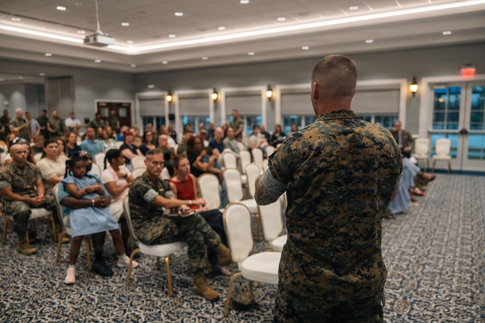 A military personnel in U.S. Marine Corps MARPAT camouflage uniform is addressing a mixed audience of uniformed Marines and civilians in a formal banquet or conference room. The speaker stands with their back to the camera, gesturing toward the seated crowd. The event appears to be an official briefing, town hall, or family readiness gathering based on the mix of military and civilian attendees.