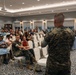 A military personnel in U.S. Marine Corps MARPAT camouflage uniform is addressing a mixed audience of uniformed Marines and civilians in a formal banquet or conference room. The speaker stands with their back to the camera, gesturing toward the seated crowd. The event appears to be an official briefing, town hall, or family readiness gathering based on the mix of military and civilian attendees.