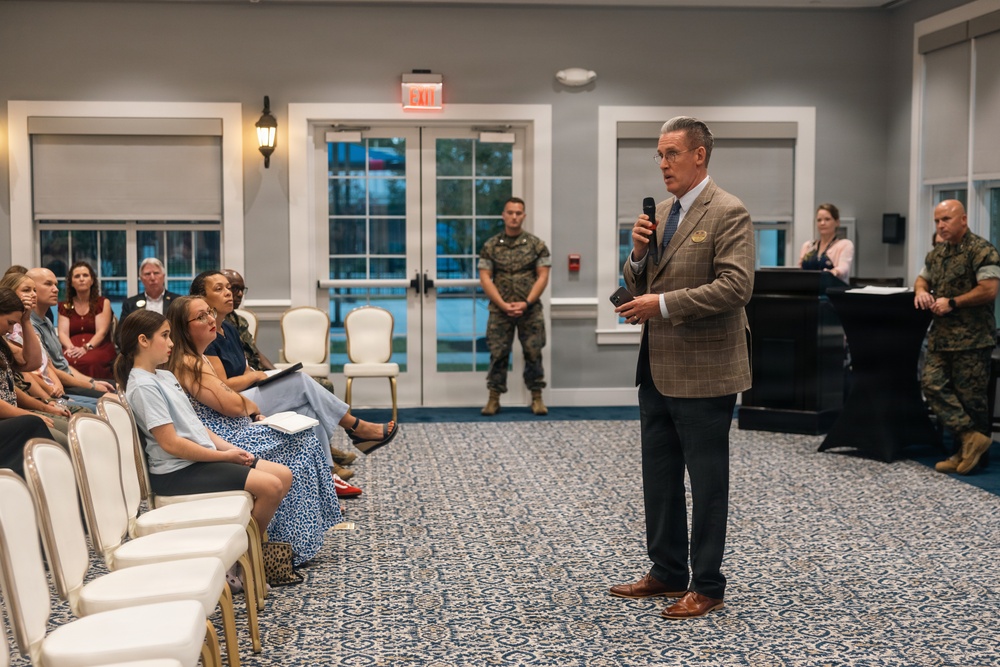 A formal indoor gathering or briefing is taking place in what appears to be a meeting hall or event room. A civilian man in a tan plaid blazer is speaking into a microphone while addressing a seated audience, with several military personnel in camouflage uniforms present in the background. The setting suggests an official military-community or town hall-style event.