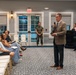 A formal indoor gathering or briefing is taking place in what appears to be a meeting hall or event room. A civilian man in a tan plaid blazer is speaking into a microphone while addressing a seated audience, with several military personnel in camouflage uniforms present in the background. The setting suggests an official military-community or town hall-style event.