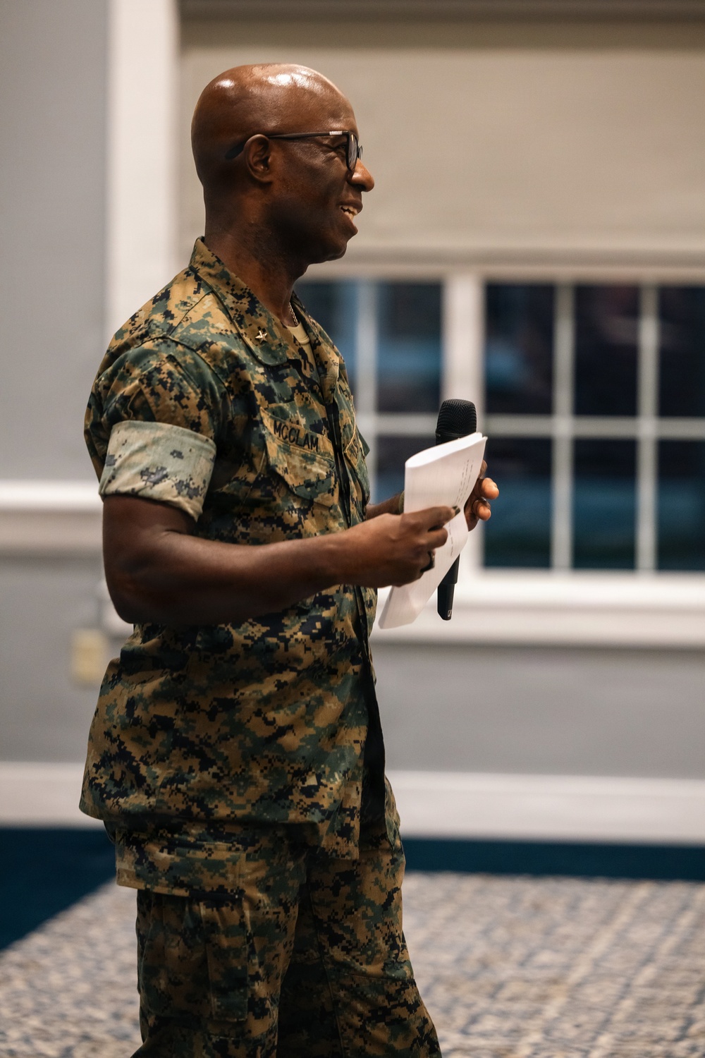A U.S. Marine Corps officer wearing MARPAT digital camouflage uniform is speaking at an indoor event, holding a microphone and papers. The name tape on the uniform reads 'MCCLAM' and a single gold star insignia is visible on the collar, indicating the rank of Brigadier General. The individual is smiling and appears to be addressing an audience in what looks like a conference or event hall.