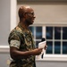 A U.S. Marine Corps officer wearing MARPAT digital camouflage uniform is speaking at an indoor event, holding a microphone and papers. The name tape on the uniform reads 'MCCLAM' and a single gold star insignia is visible on the collar, indicating the rank of Brigadier General. The individual is smiling and appears to be addressing an audience in what looks like a conference or event hall.