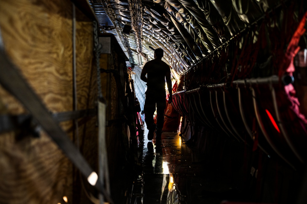 A silhouetted figure walks through the interior cargo hold of a large military transport aircraft, likely a C-17 or C-130. The curved fuselage walls with ribbed metallic framing are visible on both sides, along with cargo straps, red seating or cargo netting, and a reflective wet floor. The dramatic backlighting creates a strong silhouette effect with a light source visible at the far end of the fuselage.
