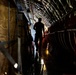 A silhouetted figure walks through the interior cargo hold of a large military transport aircraft, likely a C-17 or C-130. The curved fuselage walls with ribbed metallic framing are visible on both sides, along with cargo straps, red seating or cargo netting, and a reflective wet floor. The dramatic backlighting creates a strong silhouette effect with a light source visible at the far end of the fuselage.