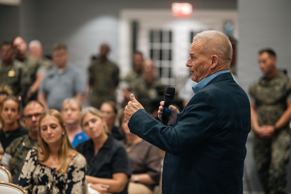 An elderly man in a dark blazer and blue shirt is speaking into a handheld microphone, addressing a mixed audience of civilians and military personnel in camouflage uniforms. The setting appears to be an indoor event or briefing room, with attendees seated and standing in the background. The speaker is gesturing with his hand, suggesting an engaging or emphatic presentation.