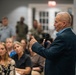 An elderly man in a dark blazer and blue shirt is speaking into a handheld microphone, addressing a mixed audience of civilians and military personnel in camouflage uniforms. The setting appears to be an indoor event or briefing room, with attendees seated and standing in the background. The speaker is gesturing with his hand, suggesting an engaging or emphatic presentation.