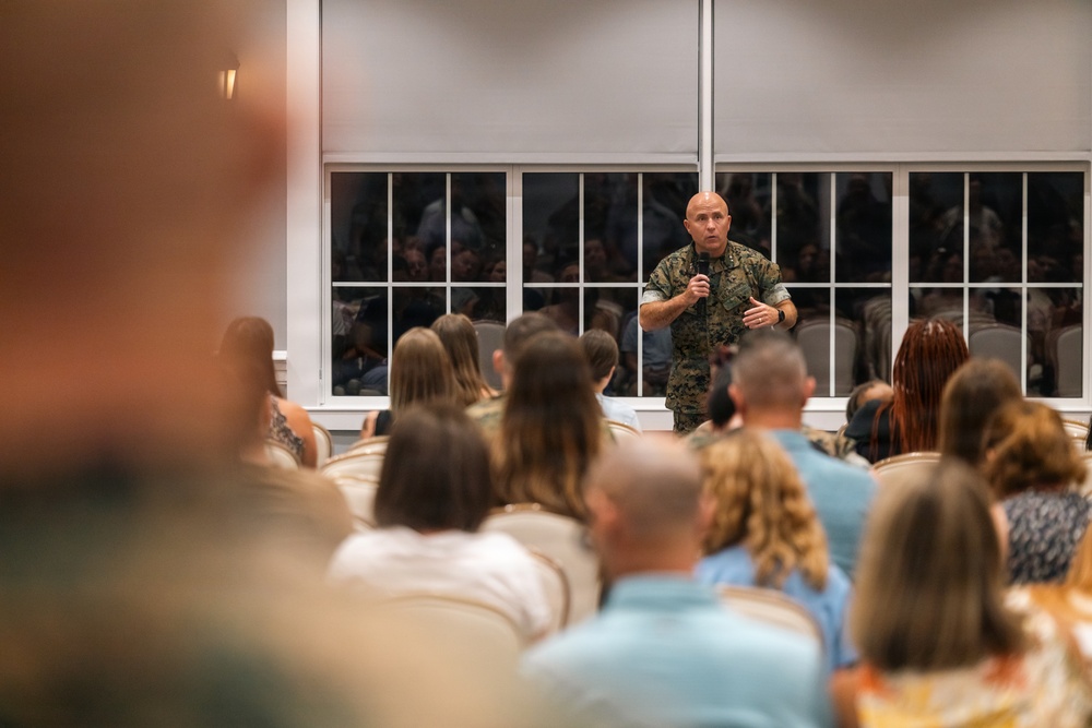 A military officer in camouflage uniform is addressing a seated civilian audience in what appears to be a briefing or town hall meeting. The speaker is holding a microphone and gesturing while speaking to a large group of attendees seated in rows. The audience appears to consist primarily of civilians, possibly military family members or community members.