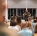 A military officer in camouflage uniform is addressing a seated civilian audience in what appears to be a briefing or town hall meeting. The speaker is holding a microphone and gesturing while speaking to a large group of attendees seated in rows. The audience appears to consist primarily of civilians, possibly military family members or community members.
